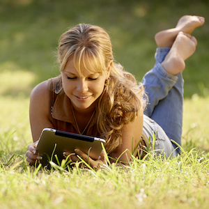 Barefoot young woman outstretched on grass smiling at computer tablet with her backpack close by
