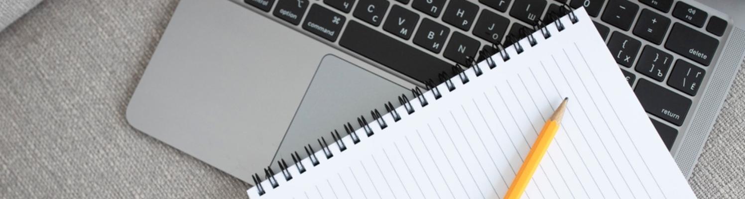 A pencil rests on a spiral-bound notebook. The notebook rests on a laptop keyboard.
