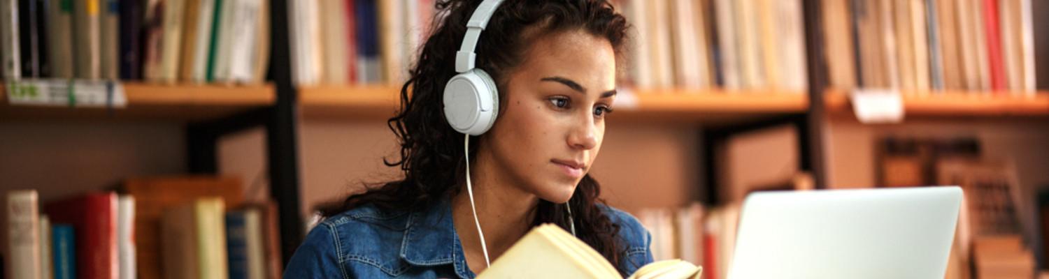 Young woman wearing headphones reading a book and browsing on a laptop