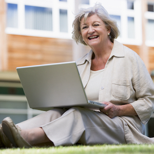 Older woman smiling while sitting on grass and using a laptop computer