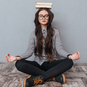 Young woman meditating cross-legged on the floor with  eyes closed and two books  balancing on her head before a gray background