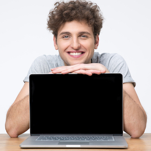 smiling young man with curly hair sitting behind an open laptop computer and learning gently on the top of the screen
