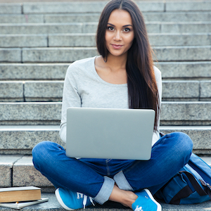 young woman with long hair and blue tennis shoes sitting on stairs and using a laptop computer