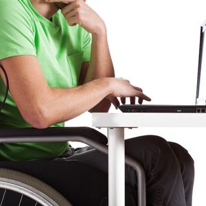 person in green shirt sitting in a wheelchair with their left hand on their chin and their right hand working on a computer keyboard