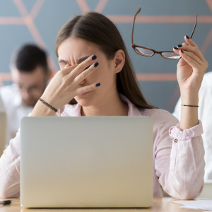 Woman sitting at a laptop pulls off her glasses and rubs the bridge of her nose, apparently under stress