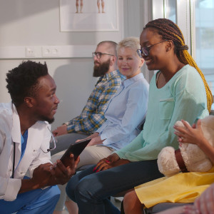 A Black doctor kneels to speak to a Black mother and her daughter in a waiting room