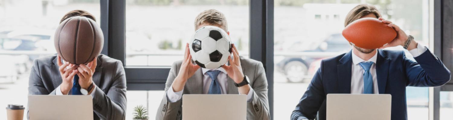 Three men sit behind laptops and hold a basketball, a soccer ball, and a football before their faces