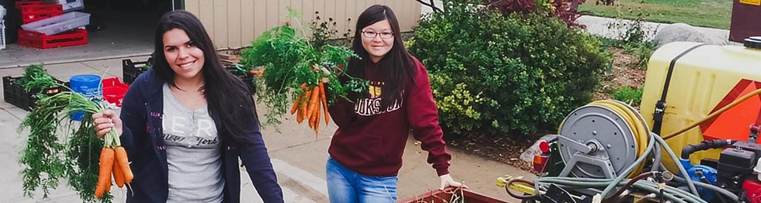 Two students pose by a tractor holding bunches of carrots