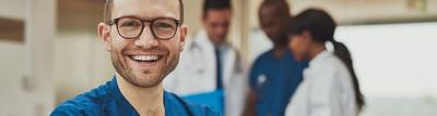 Smiling young nurse standing with his arms folded in front of some of his colleagues who are engaged in conversation